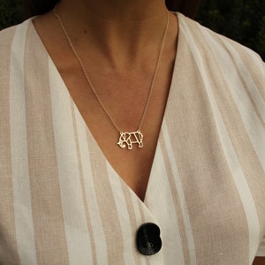 May include: A gold-colored necklace featuring a geometric rhino pendant. The rhino pendant is suspended from a delicate chain. The necklace is worn over a beige and white striped top with a black button.
