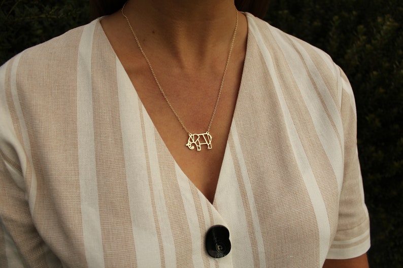 May include: A gold-colored necklace featuring a geometric rhino pendant. The rhino pendant is suspended from a delicate chain. The necklace is worn over a beige and white striped top with a black button.