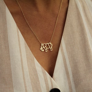 May include: A gold-colored necklace featuring a geometric elephant pendant. The pendant is a stylized representation of an elephant, with a delicate chain. The necklace is worn against a striped beige and white fabric.