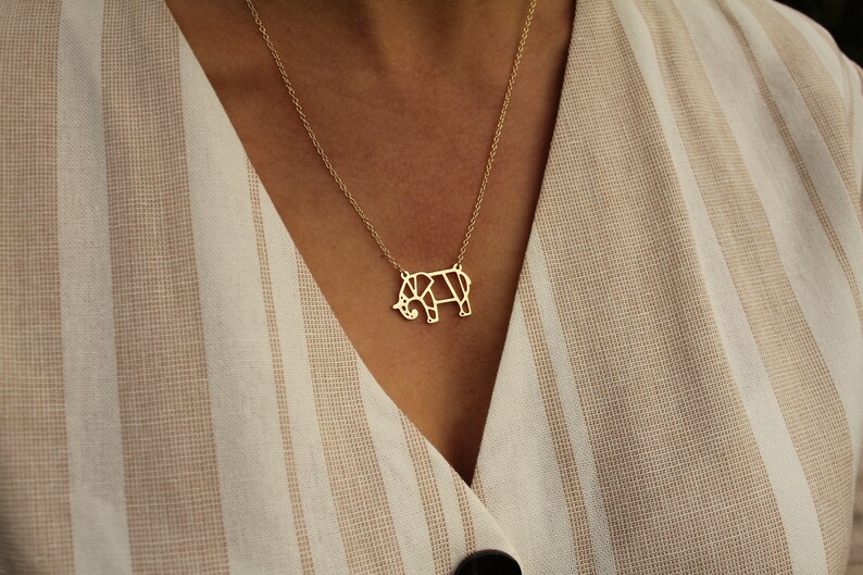 May include: A gold-colored necklace featuring a geometric elephant pendant. The pendant is a stylized representation of an elephant, with a delicate chain. The necklace is worn against a striped beige and white fabric.