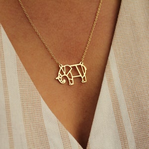 May include: A gold-colored necklace featuring a geometric elephant pendant. The elephant design is made of connected lines, creating a modern, minimalist look. The necklace is worn against a striped beige and white background.
