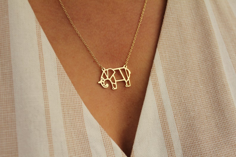 May include: A gold-colored necklace featuring a geometric elephant pendant. The elephant design is made of connected lines, creating a modern, minimalist look. The necklace is worn against a striped beige and white background.