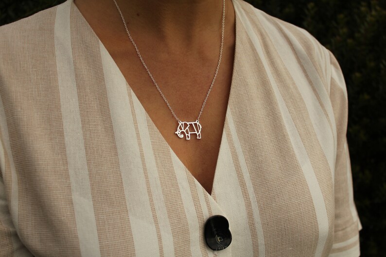May include: A silver necklace featuring a geometric elephant pendant. The pendant is a stylized outline of an elephant. The necklace is worn over a beige and white striped top.