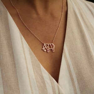 May include: Rose gold elephant pendant necklace. The necklace features a geometric elephant pendant suspended from a delicate chain. The pendant is a light rose gold color. The background is a beige and white striped fabric.