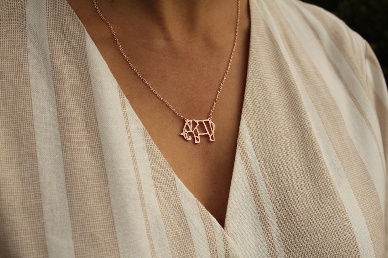 May include: Rose gold elephant pendant necklace. The necklace features a geometric elephant pendant suspended from a delicate chain. The pendant is a light rose gold color. The background is a beige and white striped fabric.