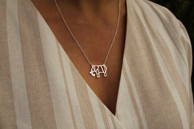 May include: A silver necklace featuring a geometric elephant pendant. The pendant is a stylized, open design with clean lines. The necklace is displayed on a person wearing a beige and white striped top.