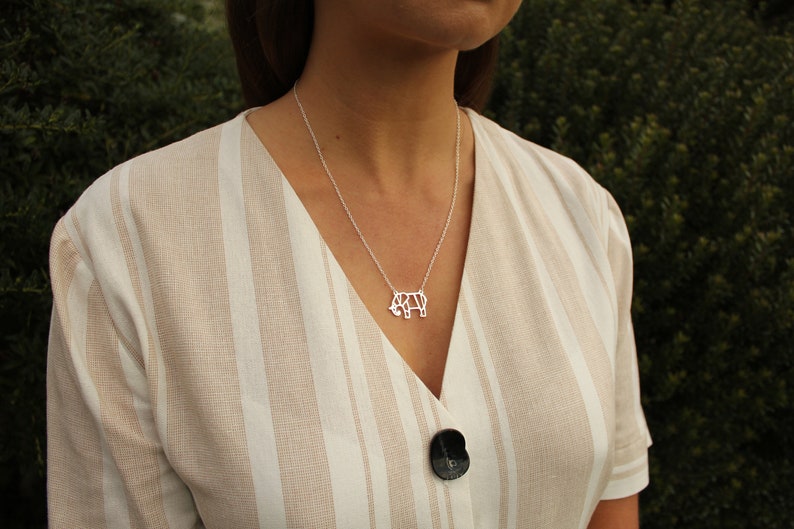 May include: A silver-colored necklace featuring a geometric rhino pendant. The necklace hangs on a delicate chain, resting on a striped beige and white dress. The background is a blurred green.