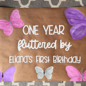 May include: A brown paper sign with the text "ONE YEAR fluttered by Eliana's FIRST BIRTHDAY" in white script. Pink and purple painted butterflies are scattered around the text, with a small gray butterfly in the center.