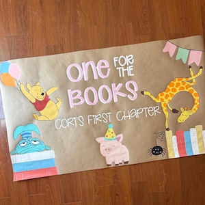 May include: A brown paper banner with the text "One for the Books" and "Cori's First Chapter" in pink. The banner features cartoon illustrations of Winnie the Pooh, a giraffe, a pig, and a fish.
