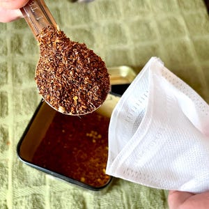 May include: A clear measuring spoon filled with loose, reddish-brown tea leaves is held above a small, open tin. A white tea bag is positioned to the right. The tea leaves are a mix of sizes and textures, suggesting a blend of ingredients.