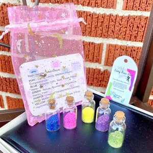 May include: Five small glass bottles filled with colorful glitter, labeled "Fairy Pixie Dust". The bottles are arranged in a row on a black surface. A pink organza bag with stars and moons printed on it is in the background. The bag contains a card with the text "Your Favorite Mood Colors" and descriptions of different colors.