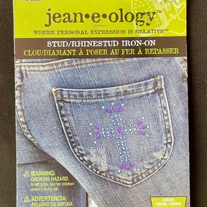 May include: A package of jean-e-ology iron-on rhinestones for clothing. The package shows a pair of blue jeans with a pocket that has a purple and silver rhinestone design. The package says "Stud/Rhinestud Iron-On" and "Clow/Diamant à poser au fer à repasser". The package also includes a warning about choking hazards for children under 3 years old.
