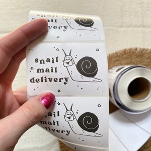 May include: A roll of white sticker labels with a black and white illustration of a snail carrying a letter. The text on the label reads "snail mail delivery".