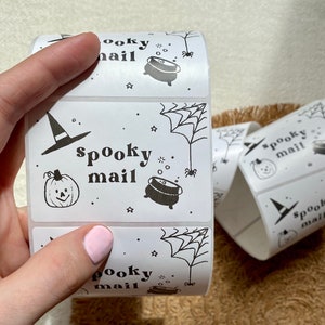 May include: Roll of white sticker labels with black and white Halloween themed illustrations. The labels feature a witch's hat, a pumpkin, a cauldron, a spider, and a spider web. The text "spooky mail" is printed on each label.