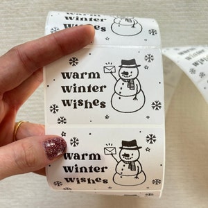 May include: A roll of white gift tags with a black and white illustration of a snowman holding a letter. The text on the tags reads "warm winter wishes".