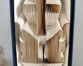 May include: A book with pages folded to create a three-dimensional sculpture of a figure. The book has a dark blue cover, and the interior pages are cream-colored with visible black printed text. The folded paper forms a central vertical shape.