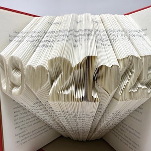 May include: A book with pages folded to display the date 09-21-25 with heart shapes between the numbers. The book has a red cover and white paper pages with black printed text. The pages are intricately creased to form the numbers and hearts.