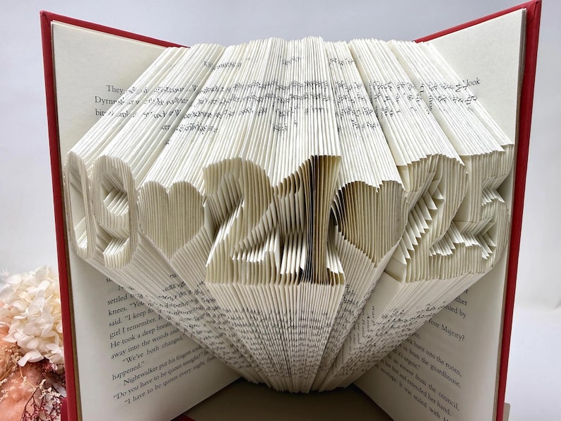 May include: A book with pages folded to display the date 09-21-25 with heart shapes between the numbers. The book has a red cover and white paper pages with black printed text. The pages are intricately creased to form the numbers and hearts.