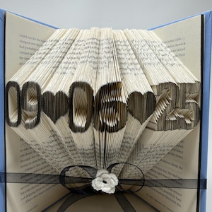 May include: A folded book art sculpture featuring the date 09-06-25 with heart shapes between the numbers. The pages are intricately folded to display the black-inked digits and symbols. A black ribbon with a small white flower is tied around the base.