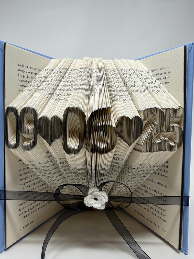 May include: A folded book art sculpture featuring the date 09-06-25 with heart shapes between the numbers. The pages are intricately folded to display the black-inked digits and symbols. A black ribbon with a small white flower is tied around the base.