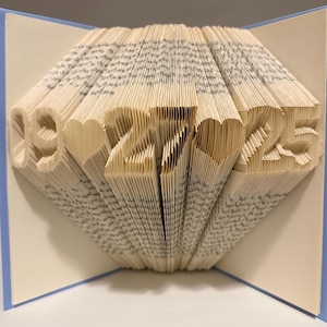 May include: A folded book art sculpture featuring the date 09-27-25 with heart shapes between the numbers. The pages are precisely creased to form the digits and hearts, set against a light blue cover that frames the white paper interior.