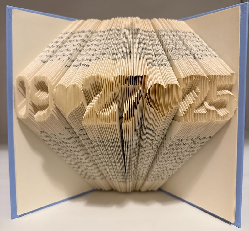May include: A folded book art sculpture featuring the date 09-27-25 with heart shapes between the numbers. The pages are precisely creased to form the digits and hearts, set against a light blue cover that frames the white paper interior.