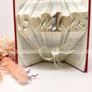 May include: A folded book art sculpture with red covers, featuring the numbers 09, 21, and 25 separated by heart shapes formed from the pages. A sheer cream-colored ribbon with a small white fabric flower is tied around the center of the book.