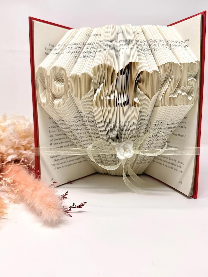 May include: A folded book art sculpture with red covers, featuring the numbers 09, 21, and 25 separated by heart shapes formed from the pages. A sheer cream-colored ribbon with a small white fabric flower is tied around the center of the book.