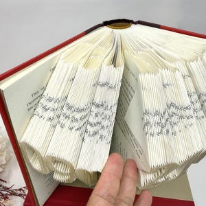May include: A hardback book with a red cover featuring pages folded to create a three-dimensional sculpture. The folded pages form the number 19, with visible printed text on the paper edges. The book is open, displaying the intricate paper art.