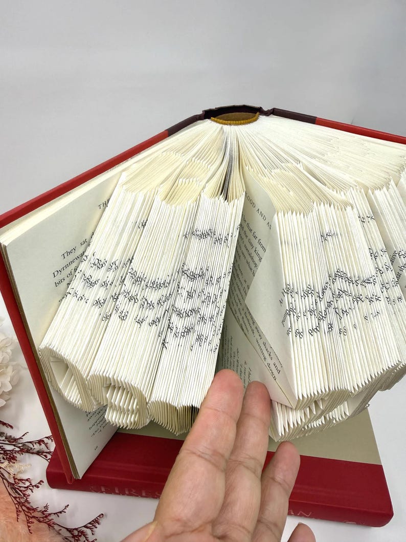 May include: A hardback book with a red cover featuring pages folded to create a three-dimensional sculpture. The folded pages form the number 19, with visible printed text on the paper edges. The book is open, displaying the intricate paper art.