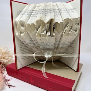 May include: A folded book art sculpture featuring the date 09-21-25 with heart symbols between the numbers. The pages are intricately folded to display the digits. A sheer ribbon with a small white flower is tied around the center of the book.