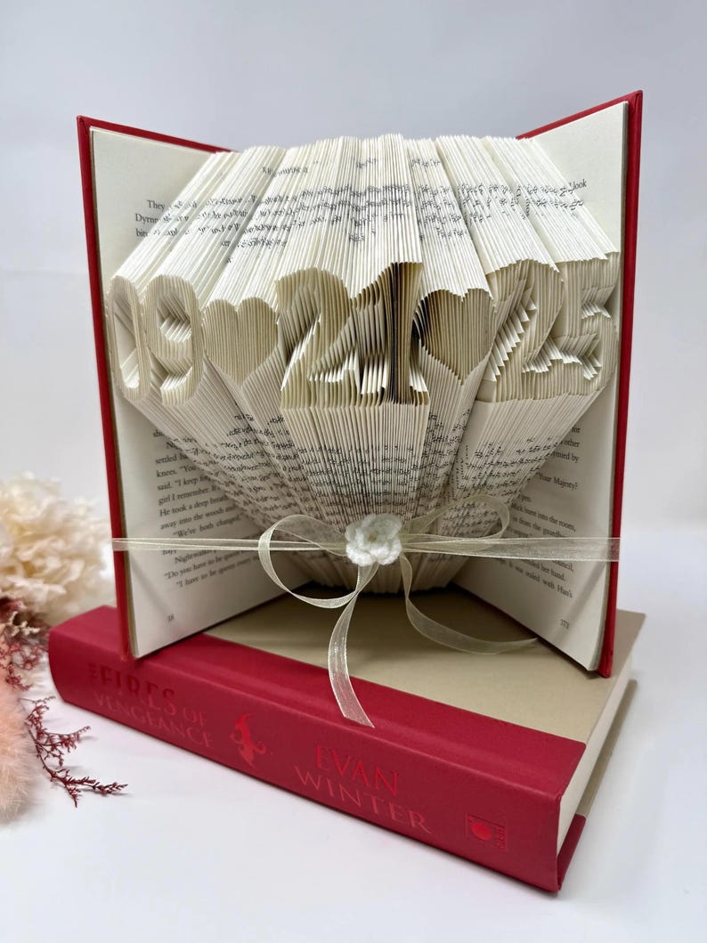 May include: A folded book art sculpture featuring the date 09-21-25 with heart symbols between the numbers. The pages are intricately folded to display the digits. A sheer ribbon with a small white flower is tied around the center of the book.
