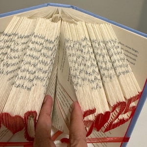 May include: A book with pages folded to create a three-dimensional design featuring red hearts and the number 25. The book has a light blue cover and white pages with black printed text. The folded edges are colored in red to highlight the shapes.