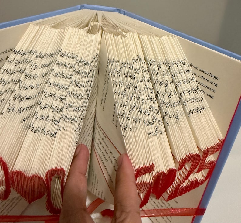 May include: A book with pages folded to create a three-dimensional design featuring red hearts and the number 25. The book has a light blue cover and white pages with black printed text. The folded edges are colored in red to highlight the shapes.