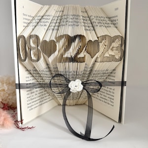 May include: A folded book art sculpture featuring the date 08-22-23 with heart shapes between the numbers. The pages are intricately creased to form the digits and symbols. A black ribbon with a small white crocheted flower is tied around it.