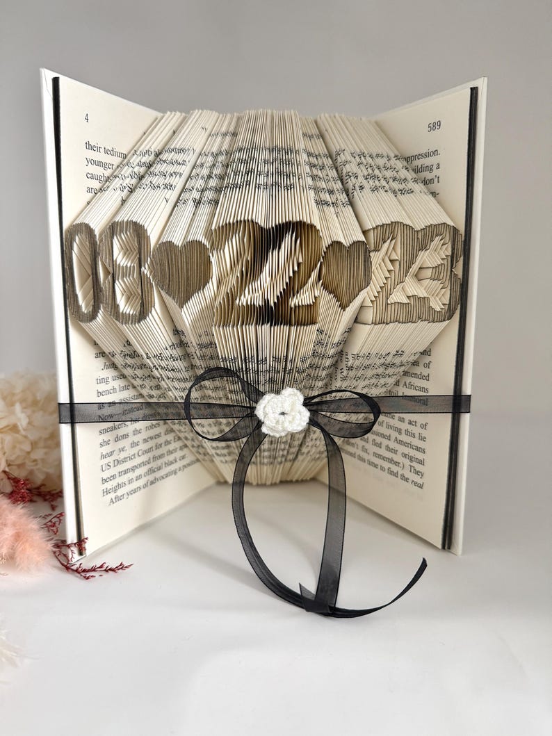 May include: A folded book art sculpture featuring the date 08-22-23 with heart shapes between the numbers. The pages are intricately creased to form the digits and symbols. A black ribbon with a small white crocheted flower is tied around it.