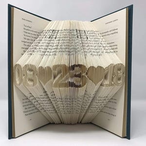 May include: A book with folded pages forming the date 08-23-18 with heart shapes between the numbers. The book has a dark blue cover and cream-colored pages with printed text visible on the open leaves. The folded pages create a 3D effect.