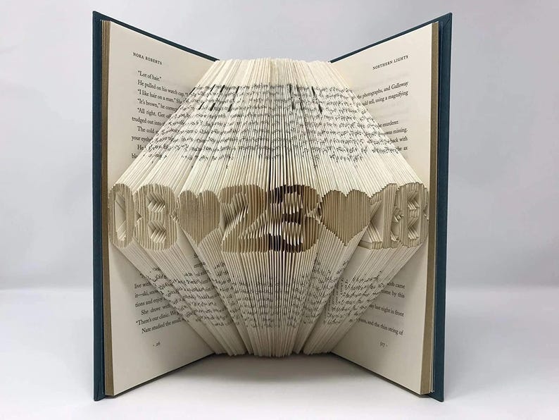 May include: A book with folded pages forming the date 08-23-18 with heart shapes between the numbers. The book has a dark blue cover and cream-colored pages with printed text visible on the open leaves. The folded pages create a 3D effect.