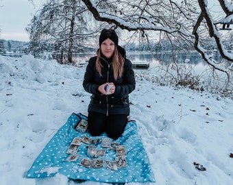 May include: A woman wearing a black hat and jacket sits on a blue and white polka dot blanket in the snow. She is holding a crystal ball and there are tarot cards spread out on the blanket.