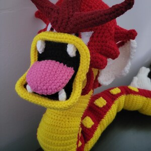 May include: A crocheted plush toy dragon with a yellow body, red and yellow stripes, and a red head with white teeth. The dragon has a pink tongue sticking out of its mouth.