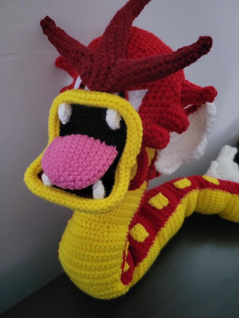 May include: A crocheted plush toy dragon with a yellow body, red and yellow stripes, and a red head with white teeth. The dragon has a pink tongue sticking out of its mouth.