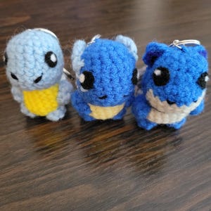 May include: Three crocheted blue and yellow keychains shaped like cartoon characters. The keychains have black eyes and are attached to silver key rings.