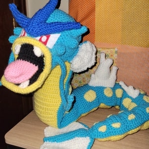 May include: A crocheted blue and yellow sea serpent with white fins and yellow spots. The serpent is curled into a circle with its mouth open.