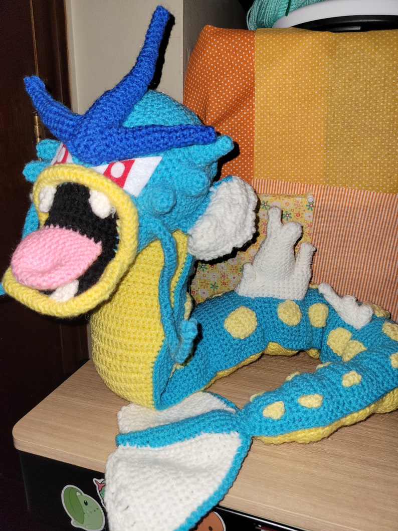May include: A crocheted blue and yellow sea serpent with white fins and yellow spots. The serpent is curled into a circle with its mouth open.