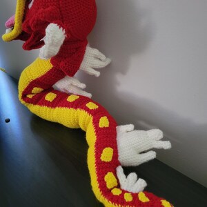 May include: A crocheted red and yellow dragon with white accents. The dragon is curled up with its head turned to the side.
