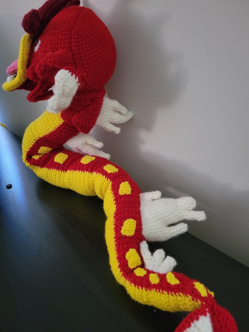 May include: A crocheted red and yellow dragon with white accents. The dragon is curled up with its head turned to the side.