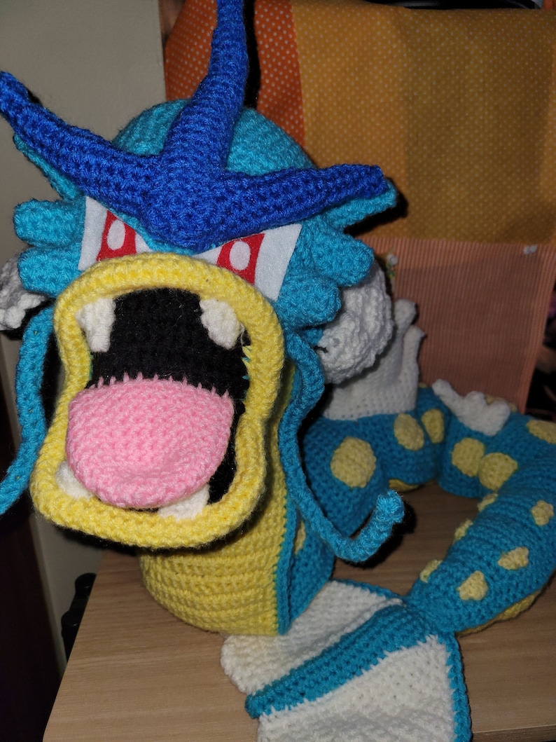 May include: A crocheted plush toy of a blue and yellow water dragon with a pink tongue. The dragon has a large, open mouth and a blue fin on its head.