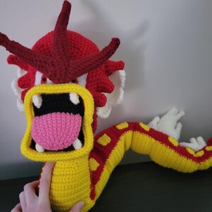 May include: A crocheted plush toy of a red and yellow dragon with a large open mouth and a pink tongue. The dragon has a long tail and is made with a variety of stitches.