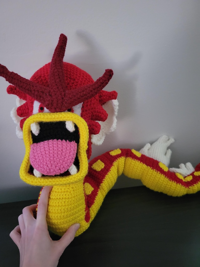 May include: A crocheted plush toy of a red and yellow dragon with a large open mouth and a pink tongue. The dragon has a long tail and is made with a variety of stitches.
