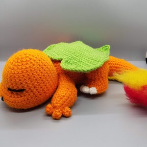 May include: A crocheted orange and green dragon plush toy with a sleeping face. The dragon has a green spiky back and a yellow and red tail.
